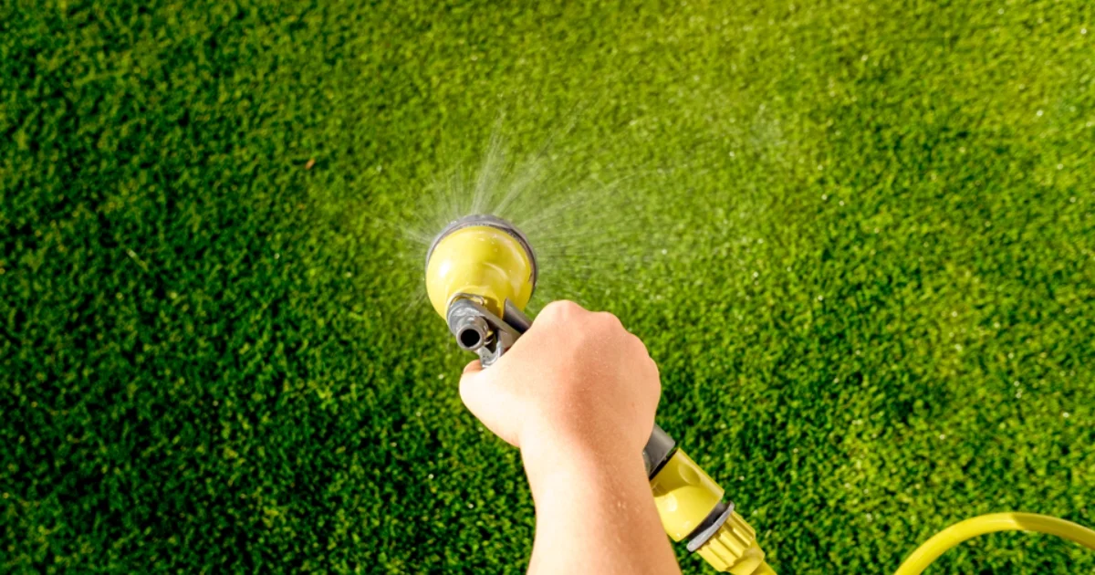  Hand holding a garden hose nozzle spraying water over synthetic turf for cleaning.